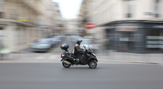 Eine Person in schwarzer Kleidung und Helm auf einem Motorroller die Straße einer Stadt entlangfahren, mit Fahrzeugen, Gebäuden und einem klaren blauen Himmel im Hintergrund.