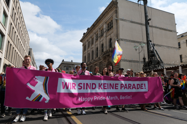 Gruppe von Menschen, die eine Straße in Berlin entlanggehen und ein pinkes "Happy Pride March"-Schild halten, mit Gebäuden auf beiden Seiten und einem Fahnenmast im Vordergrund unter einem bewölkten Himmel.