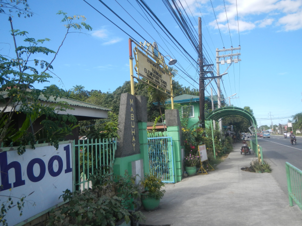 Schuleingangsschild am Straßenrand mit einem Bogen, Tor und einem Schild, umgeben von Fahrzeugen, Pflanzen, Bäumen, Strommasten und einem bewölkten Himmel.