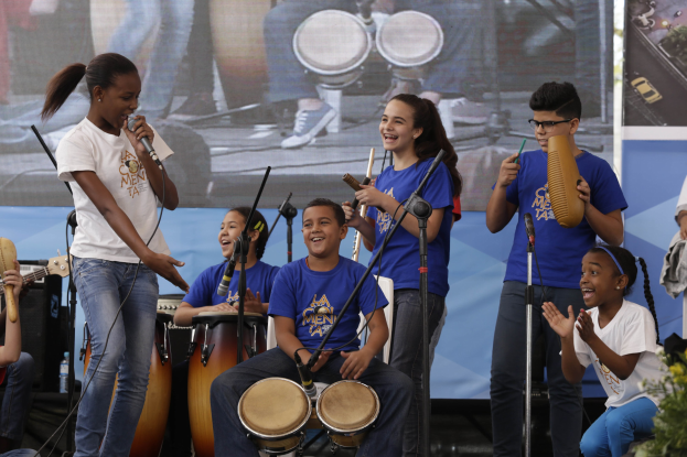 Eine Gruppe von Kindern, die auf einer Bühne Musikinstrumente spielen, mit einer Frau in der Mitte, die ein Mikrofon hält, umgeben von Mikrofonständern und einem Banner im Hintergrund, mit Pflanzen auf der rechten Seite.