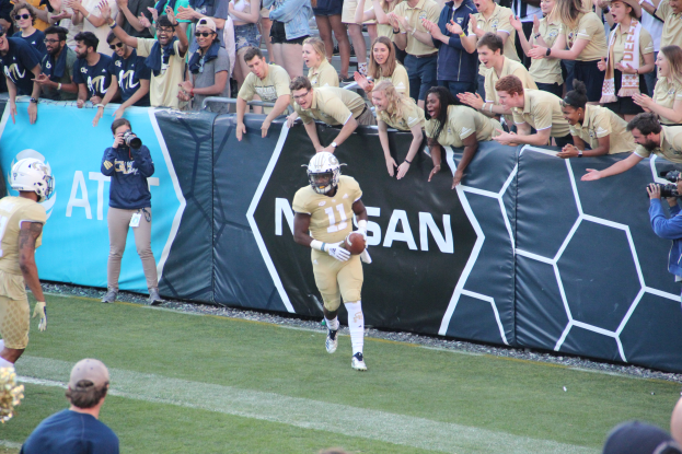 Ein Fußballspieler läuft mit dem Ball auf einem grünen Rasen vor einer Menge, einige halten Kameras und tragen Helme, mit einem Banner im Hintergrund.