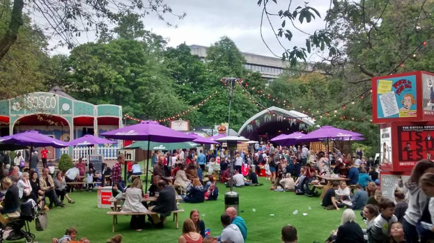 Eine große Gruppe von Menschen, die auf einem grünen Feld mit Schirmen, Tischen und einer Tafel sitzen, die ein Biergarten in London anzeigt.