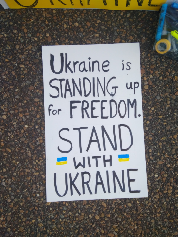 Ein weißer Zettel mit den Worten "Ukraine steht für Freiheit ein, steht mit Ukraine" liegt vor einer gelben Fahne auf dem Boden. Rechts daneben befindet sich eine Plastikhülle mit verschiedenen Gegenständen.