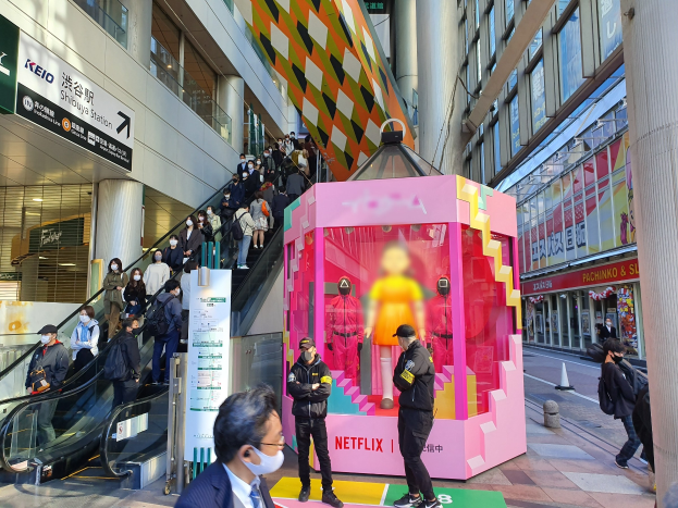 Eine Gruppe von Menschen steht vor einem Gebäude mit Glaswänden, Säulen und Texttafeln, einige tragen Masken, während eine Rolltreppe mit Menschen im Hintergrund zu sehen ist; eine pinkfarbene Box in der Mitte zeigt Schaufensterpuppen und trägt die Aufschrift "Netflix's new Netflix booth in Tokyo."