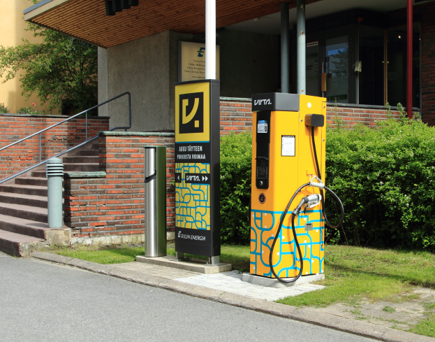 Eine Elektrofahrzeug-Ladestation an der Straße, umgeben von Grün und einem Gebäude mit Fenstern und Säulen, mit Treppen und Geländern, die hinaufführen.