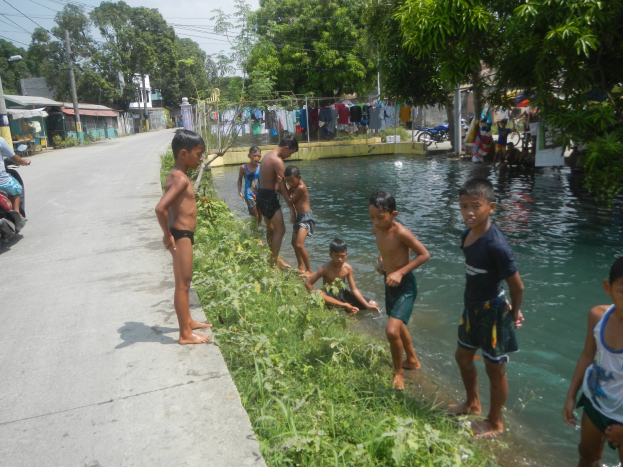 Kinder, die in flachem Wasser an der Straße spielen, einige stehen, andere sitzen auf dem Gras, mit einer Person auf einem Motorrad links und Häusern, Bäumen und einem blauen Himmel im Hintergrund.