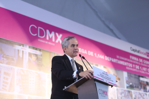 Ein Mann im Anzug und Krawatte steht hinter einem Podium mit zwei Mikrofonen und einer Tafel vor sich, mit einem Banner mit Bildern und Text dahinter.