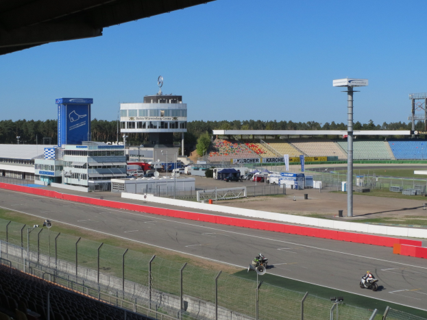 Gruppe von Motorradfahrern auf einer Rennstrecke, umgeben von einem Metallzaun, Fahrzeugen, einem Gebäude, einem Schild, Pfosten, einem Stadion, Deckenleuchten, Bäumen und einem bewölkten Himmel.