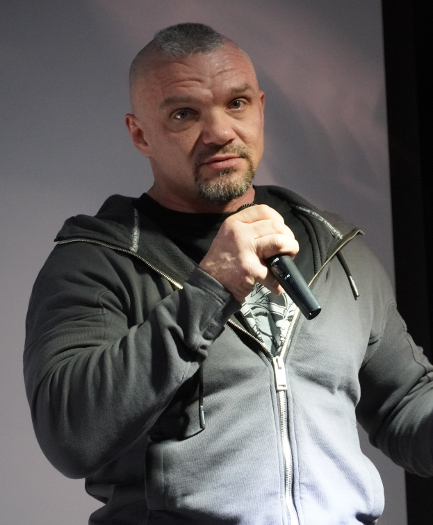 Ein Mann in einer schwarzen Kapuzenjacke und schwarzem Anzug mit einem wei├čen Hemd, der ein Mikrofon h├Ąlt und selbstbewusst vor einer Wand steht.